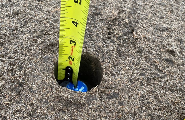 A hole in the soil with a soil temperature device and a tape measure indicating 2-inch depth