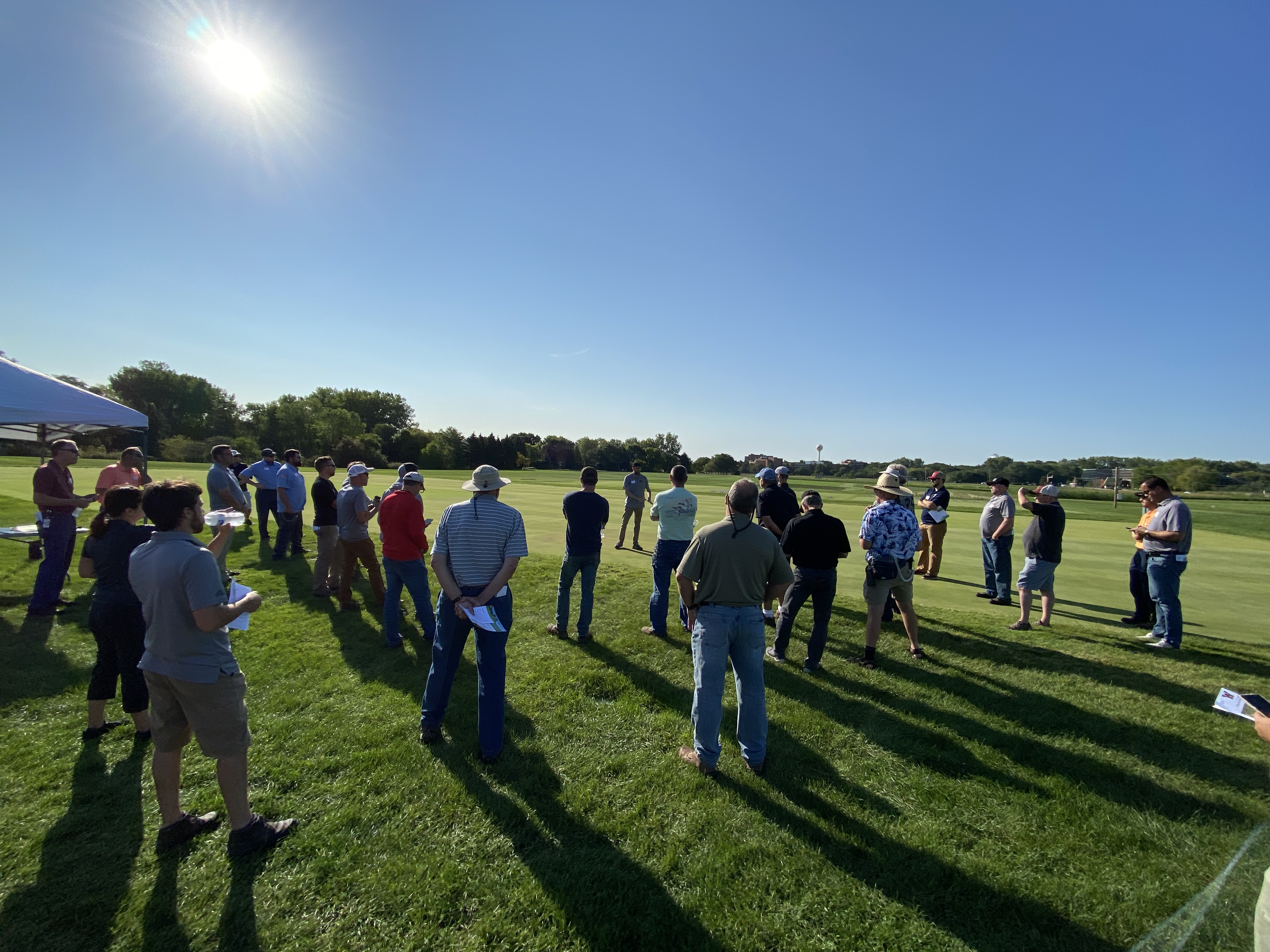a group of people listening to a speaker outside at a turf field day